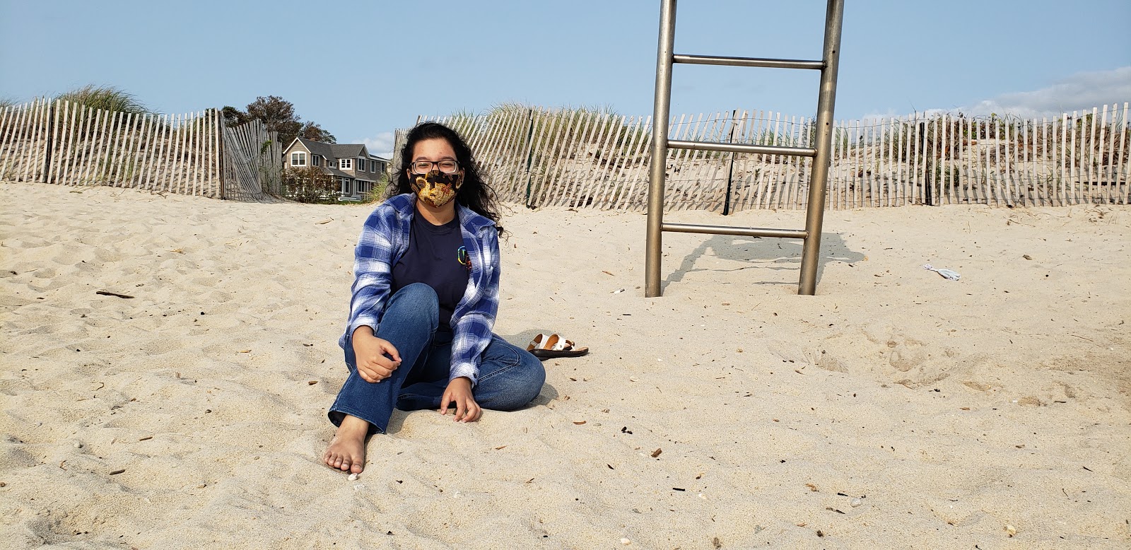 Samirah sits cross-legged in the sand at the beach with her mask on. 