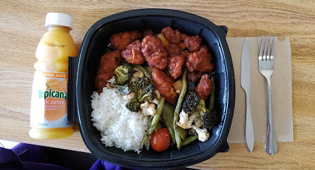 A takeout container full of veggies, white rice, and sweet and sour chicken laid out on a table with a bottle of tropicana. 