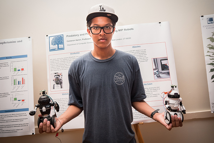 A student holds two small robots in front of a poster detailing his science research. 