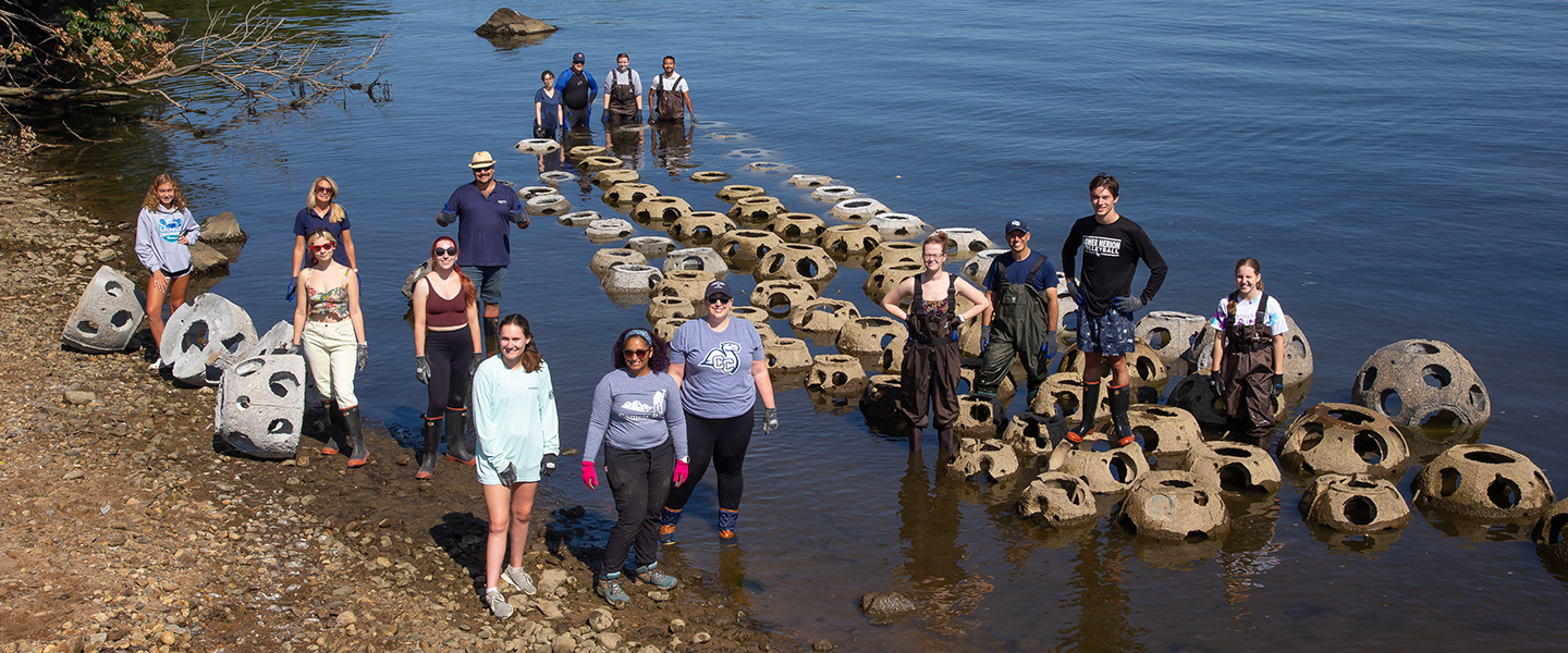 Professor Maria Rosa and her student researchers pose with the reef balls of Camels Reef, along the banks of the Thames River.