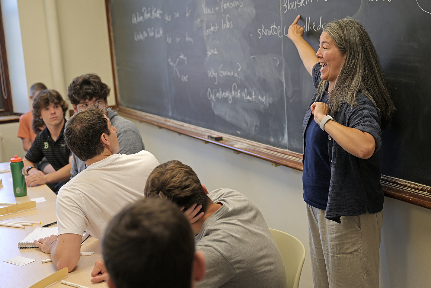 A professor smiles and points at a blackboard.