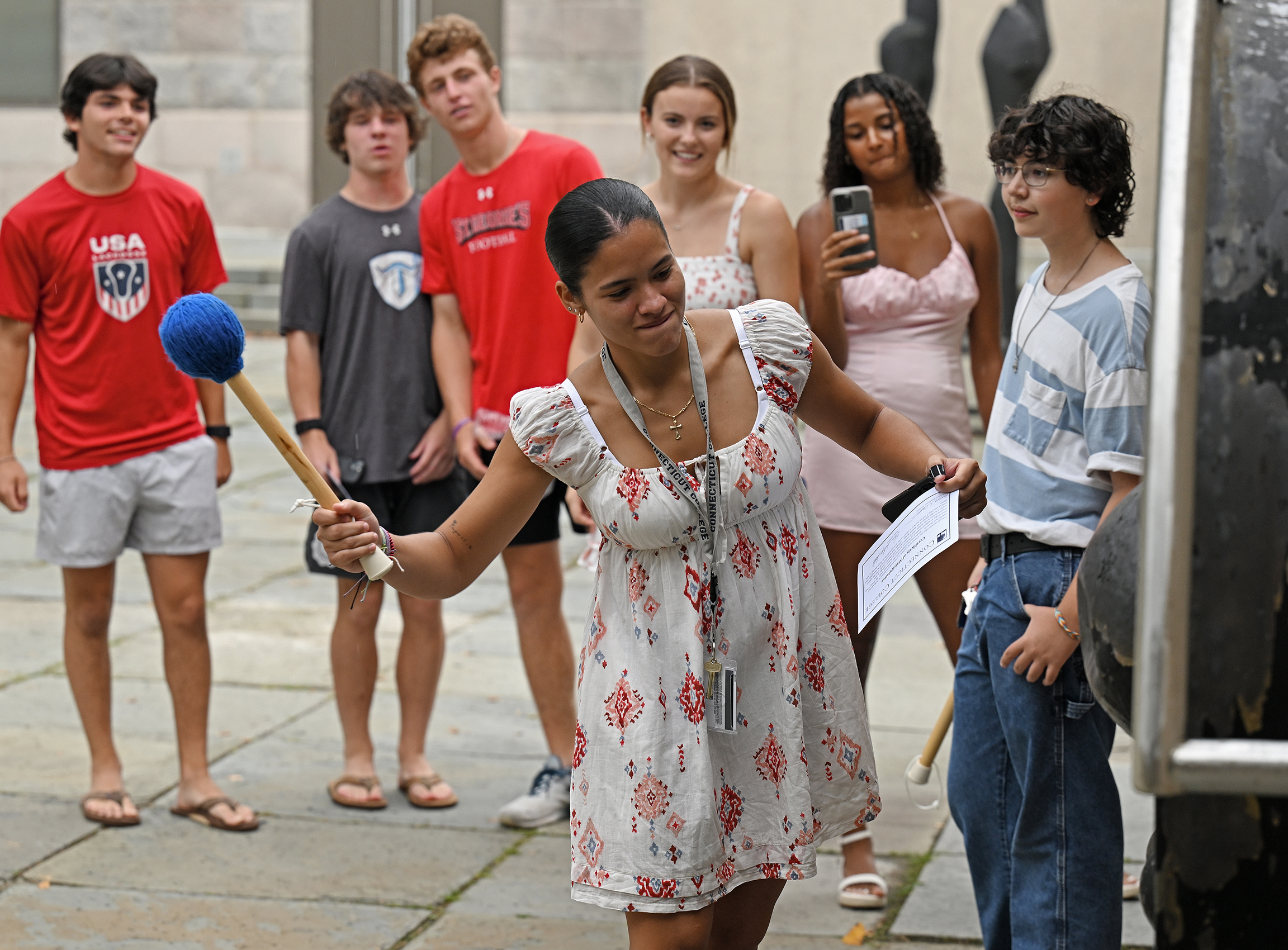 A student with a mallet bangs on a large metal gong while others watch.