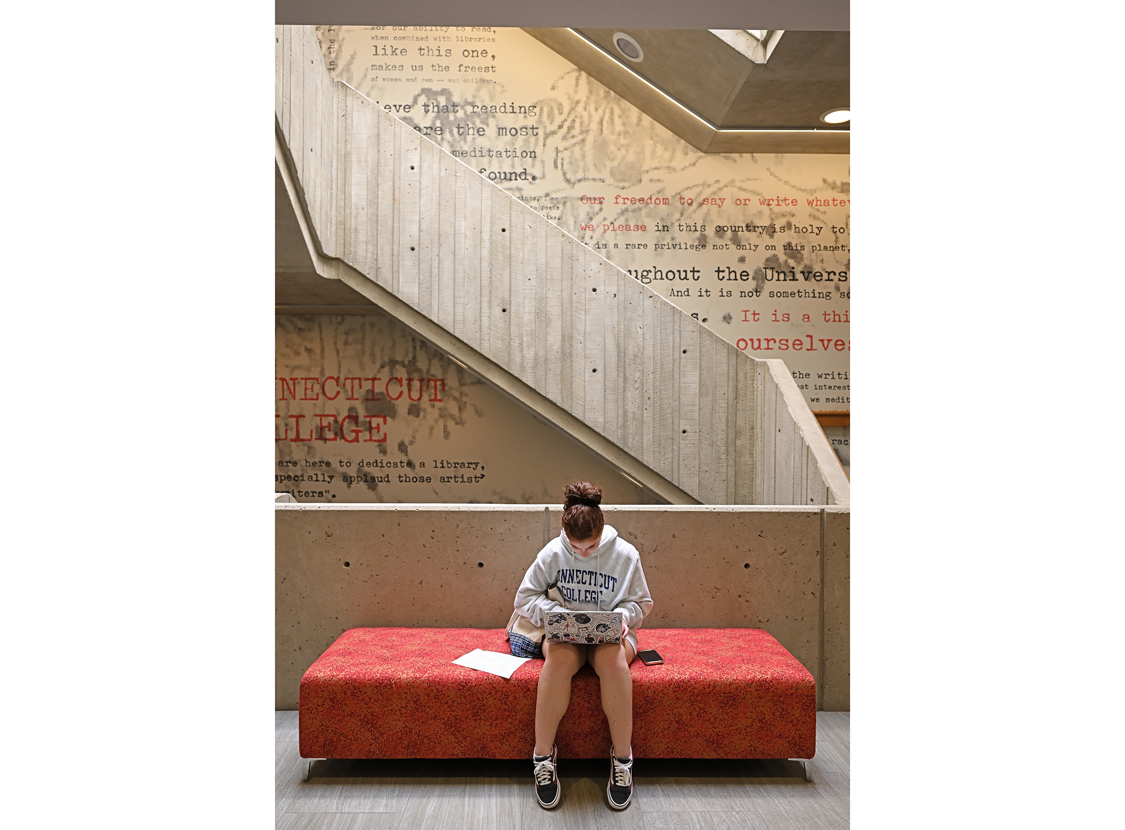 A student sits on a padded bench working on a laptop computer