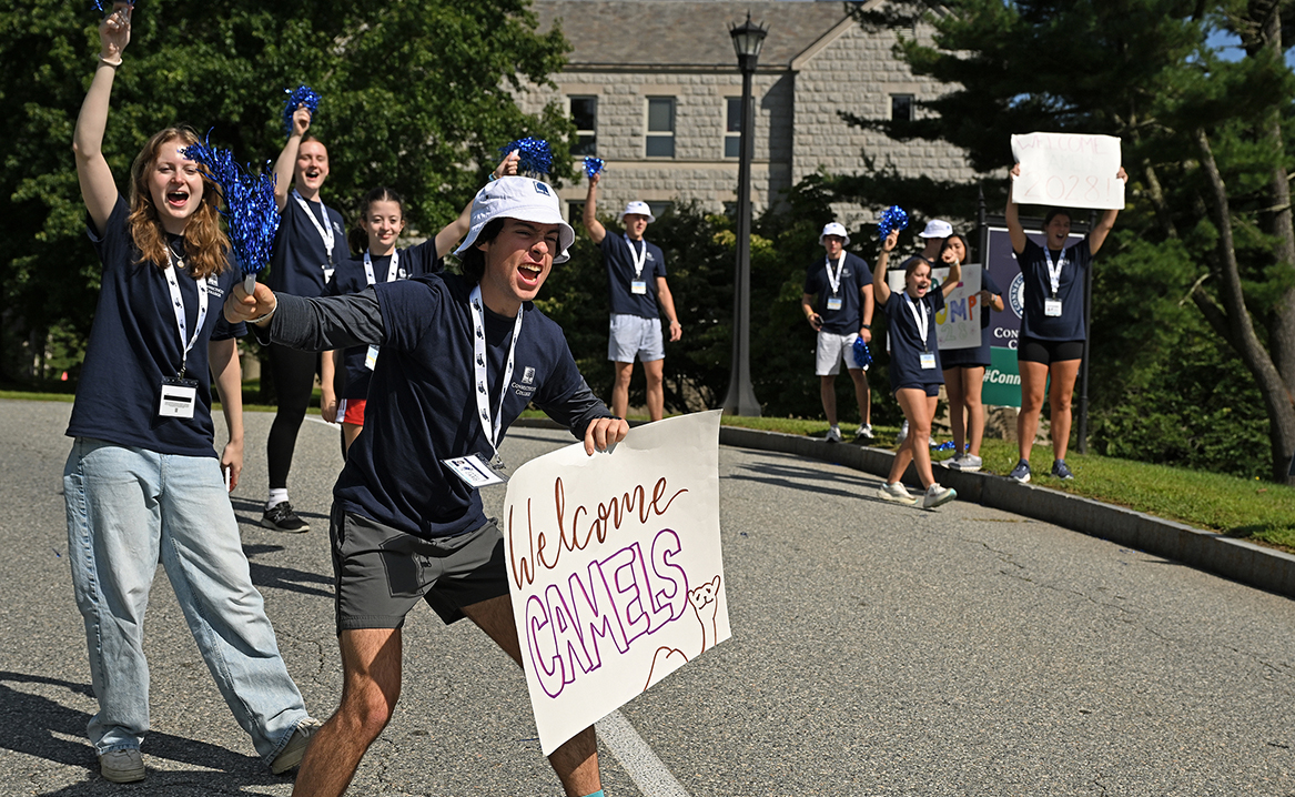 Students with handmade signs cheer and wave pom poms at a college entrance