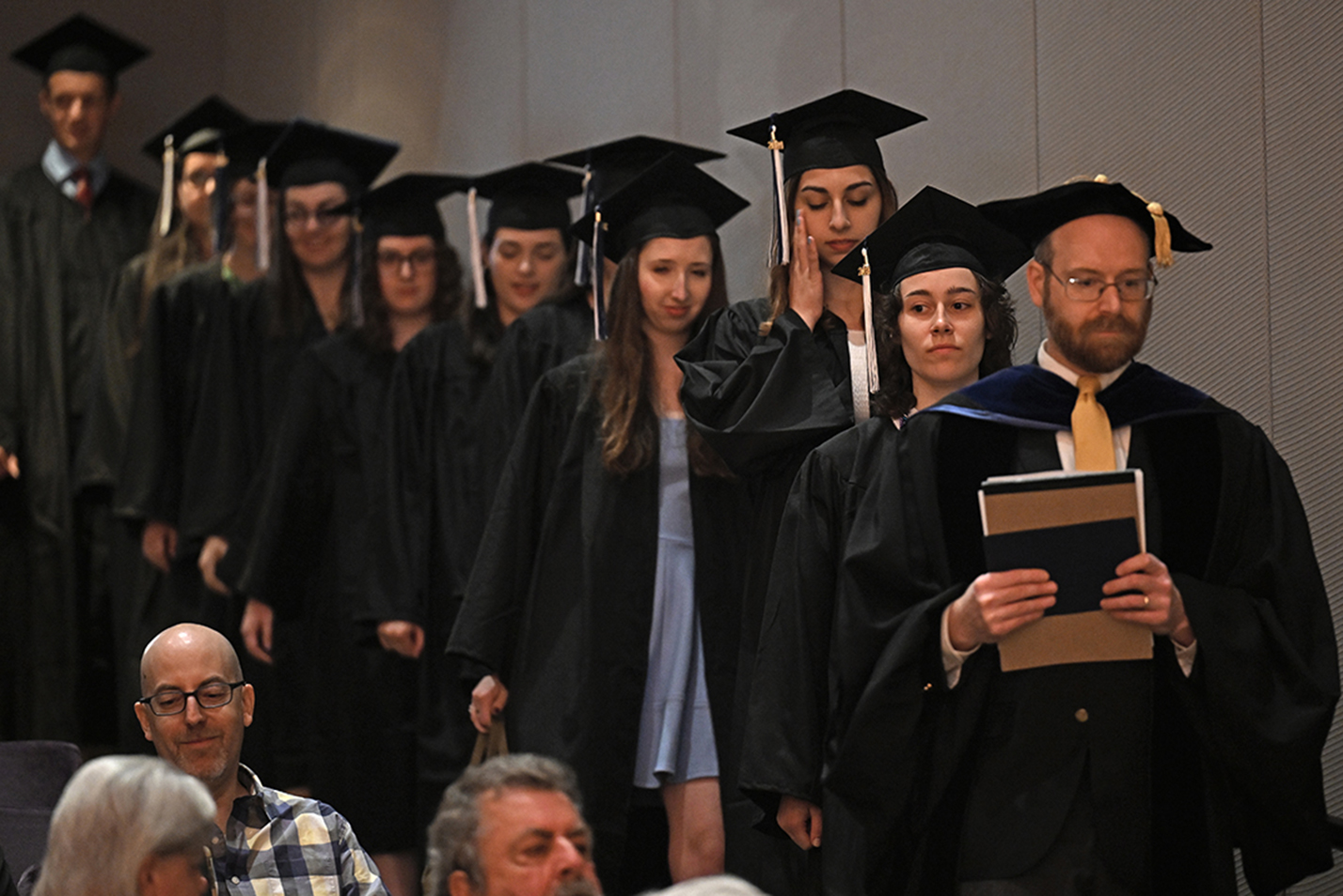 A professor leads a line of students in caps and gowns into a room for an honor society induction ceremony