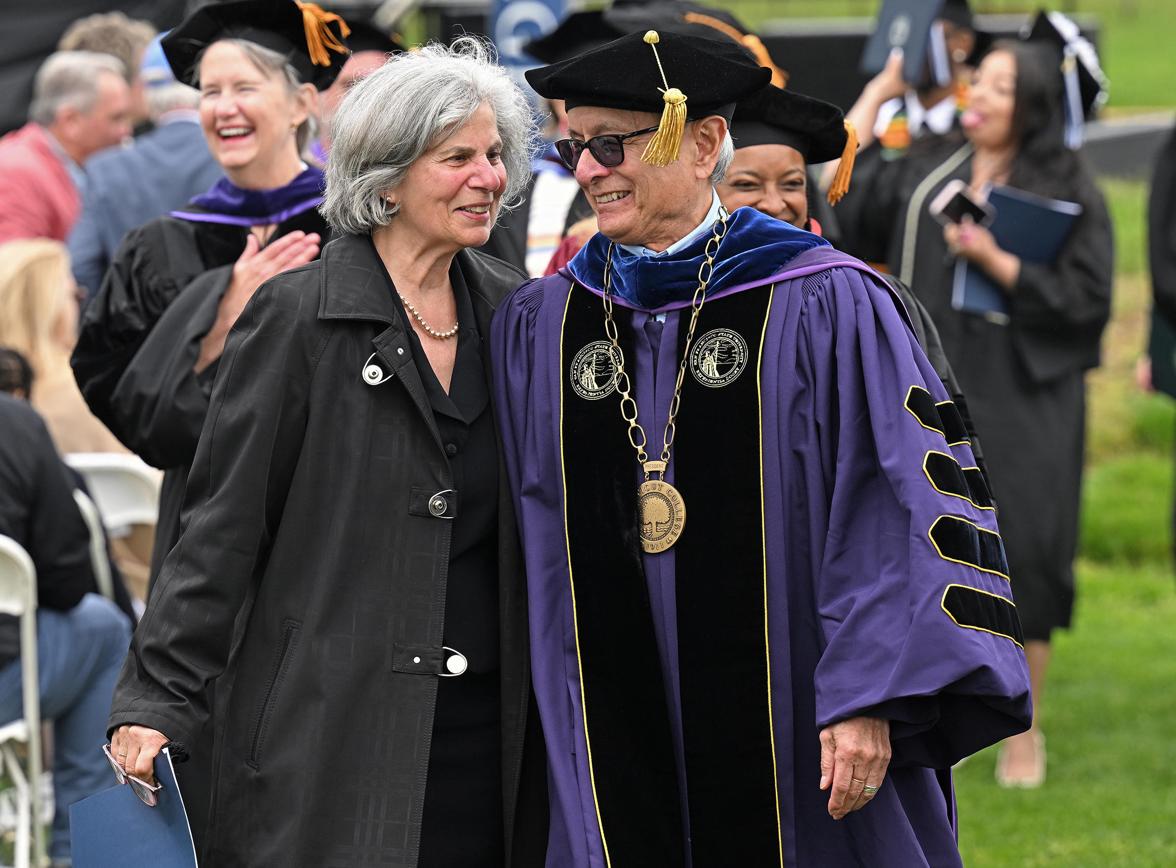 A college president in full academic regalia and his wife walk off the field together following commencement exercises