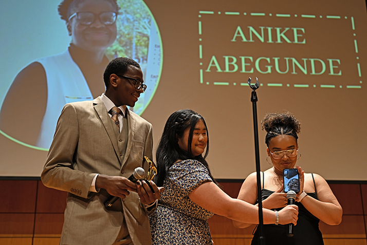 three students, one with phone in hand, stand at microphone to accept award