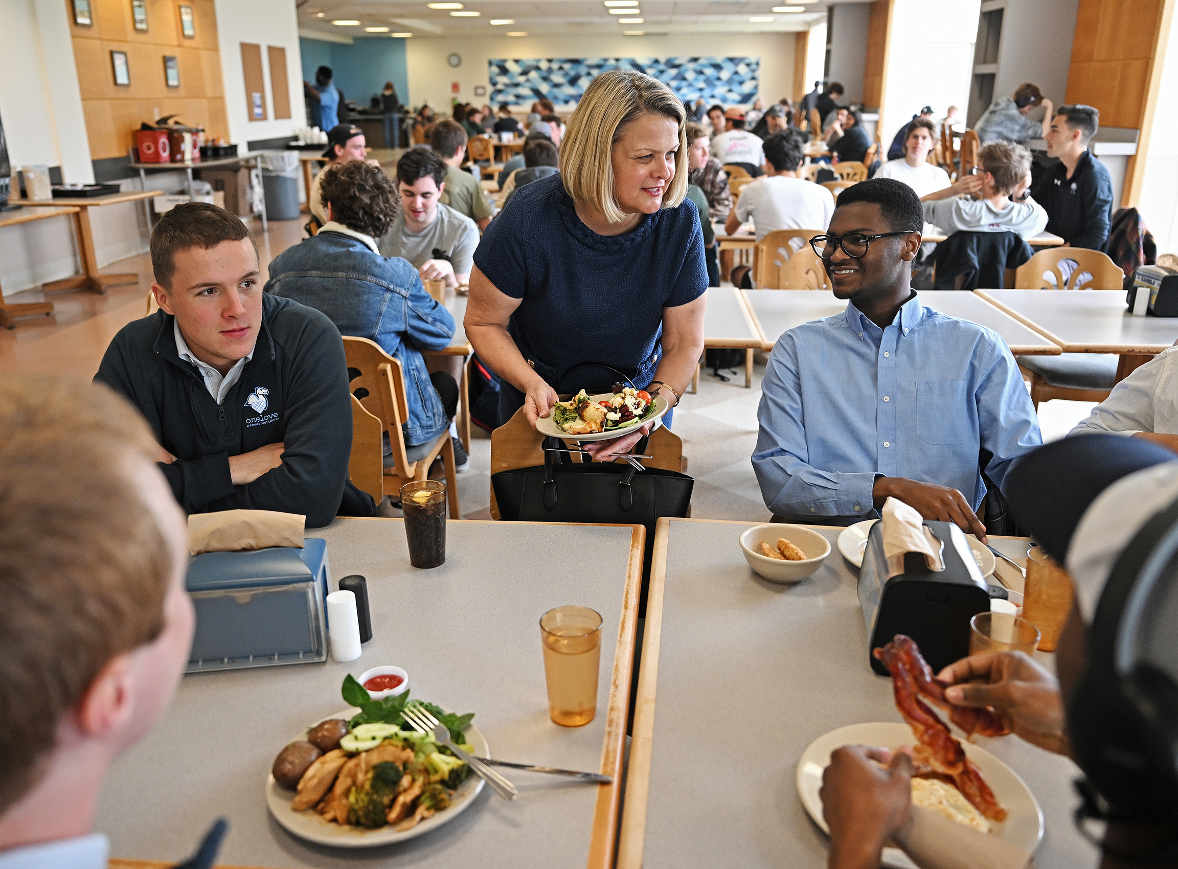 A woman in a blue dress joins a group of students at a dining hall table