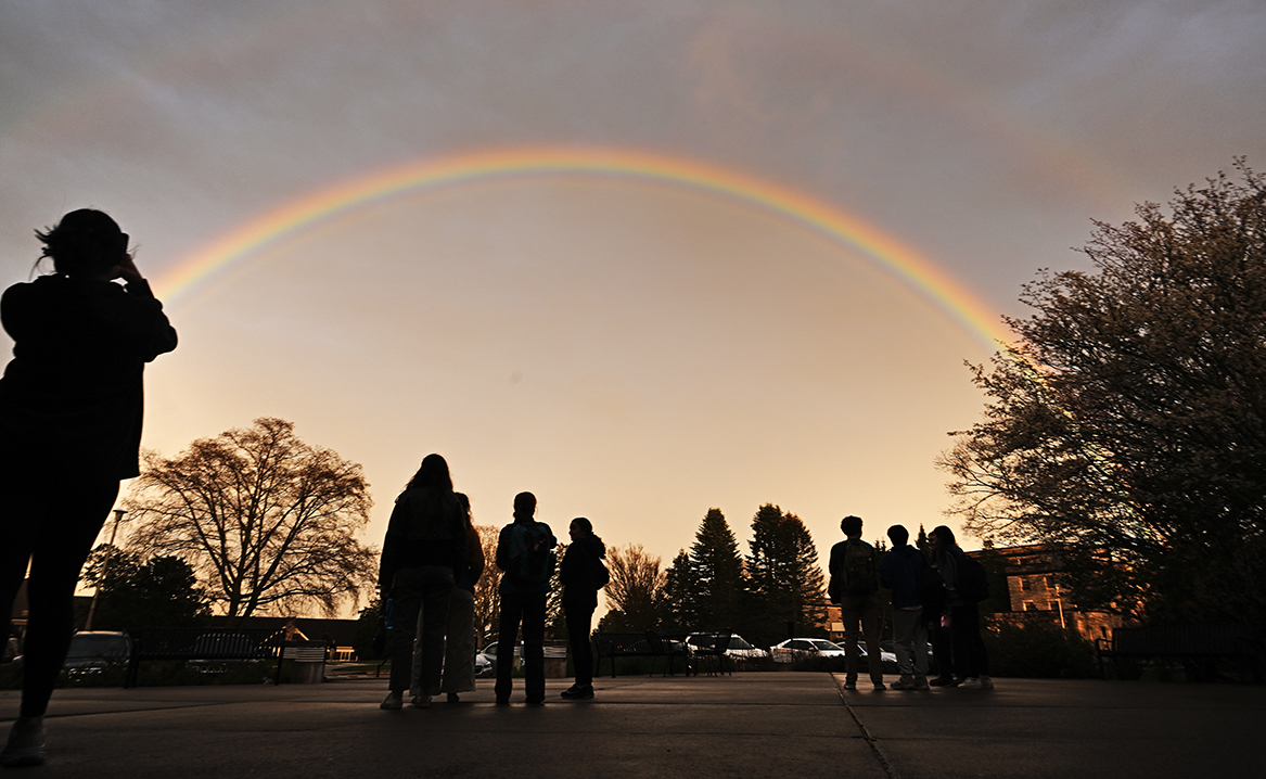 A full arc rainbow shines in the sky over a crowd gathered to view