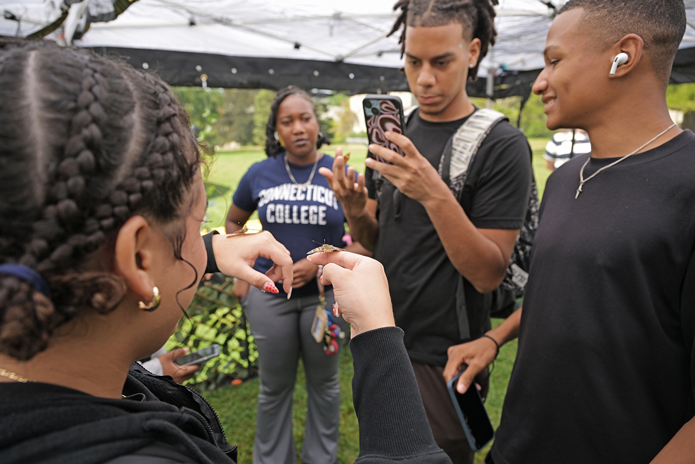 A group of students gather around another with butterflies on her hand.